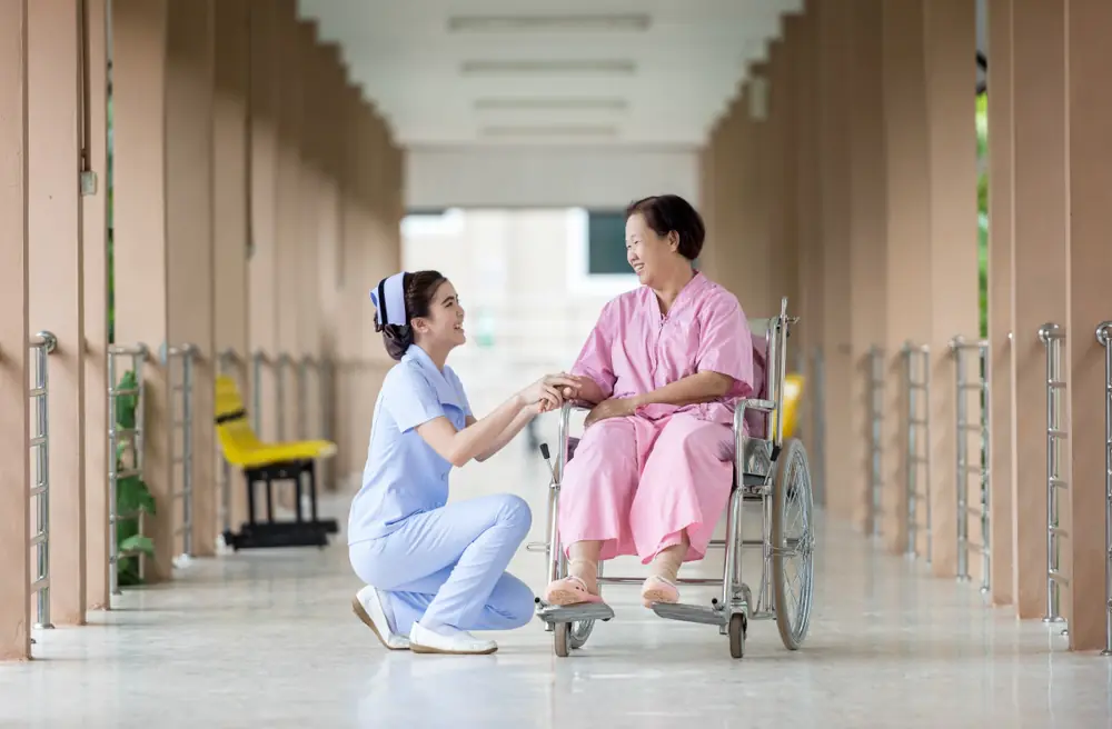 A nurse kneeling beside a patient in a wheelchair in a hospital corridor